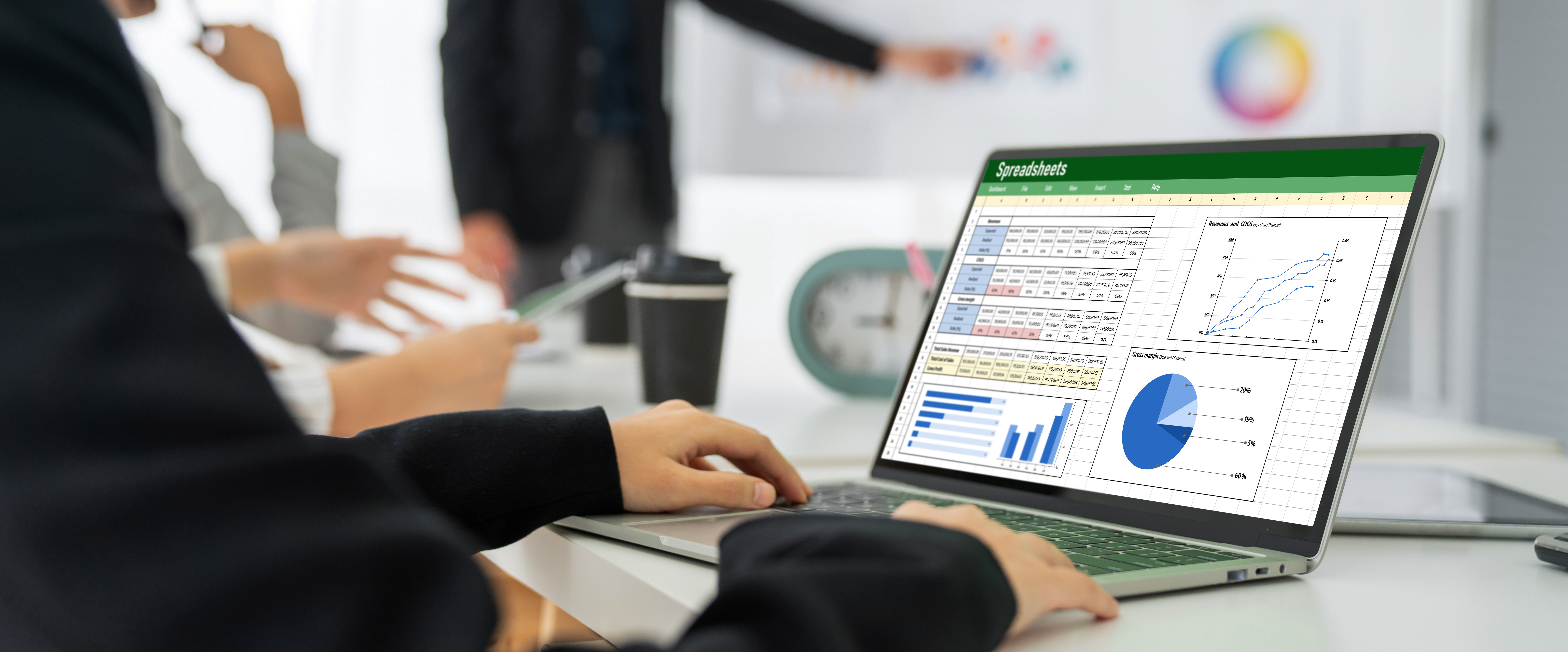 Person using a laptop displaying spreadsheets in a meeting room. Blurred colleagues and whiteboard in the background. Mood: focused.