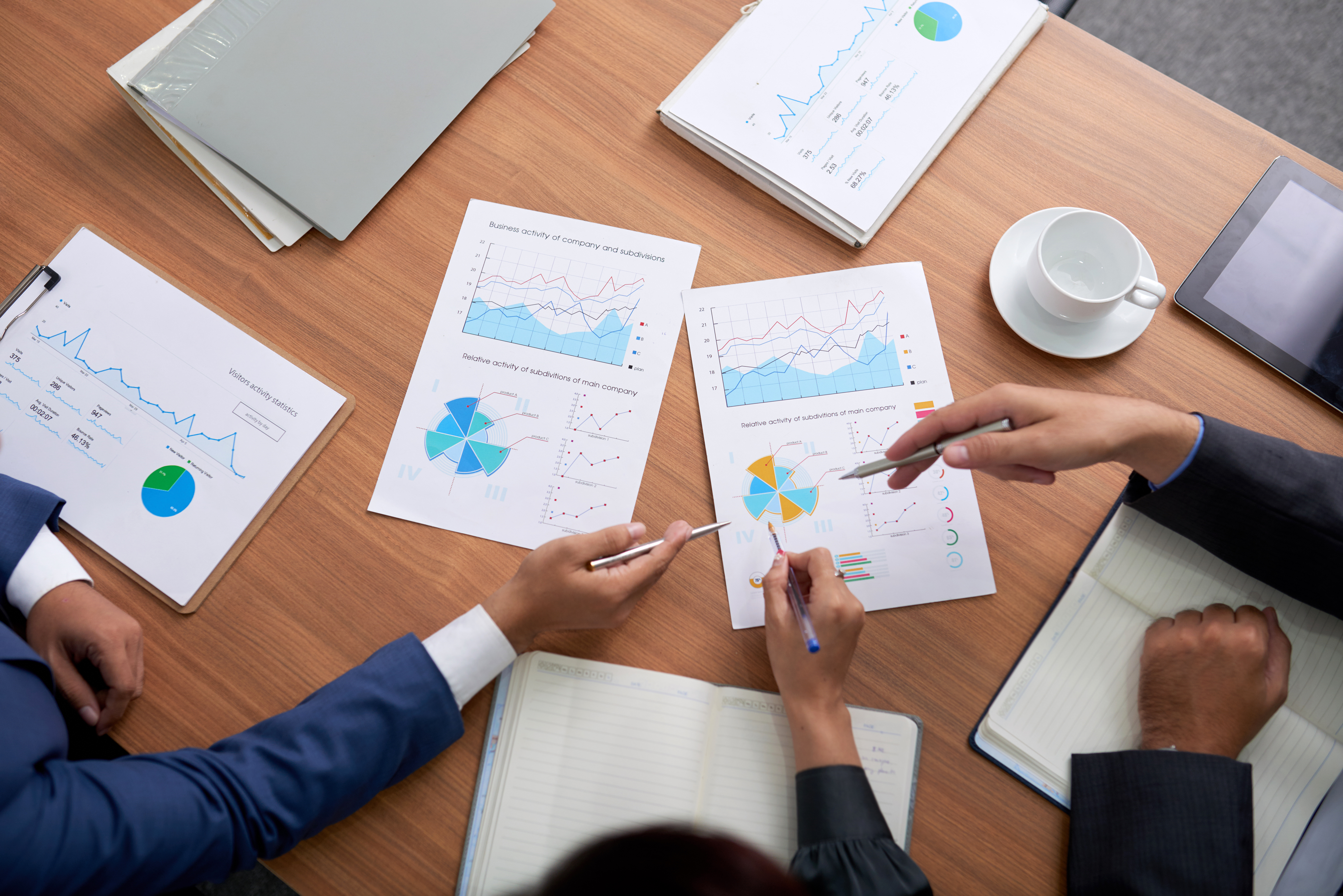 Hands reviewing colorful charts and graphs on a wooden table. Includes notebooks, a tablet, and a coffee cup. Business meeting setting.